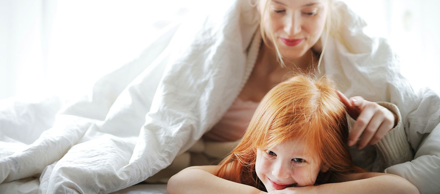 una madre en la cama justo a su hija tumbada boca abajo en la cama donde la madre le toca el pelo de forma tierna a la niña