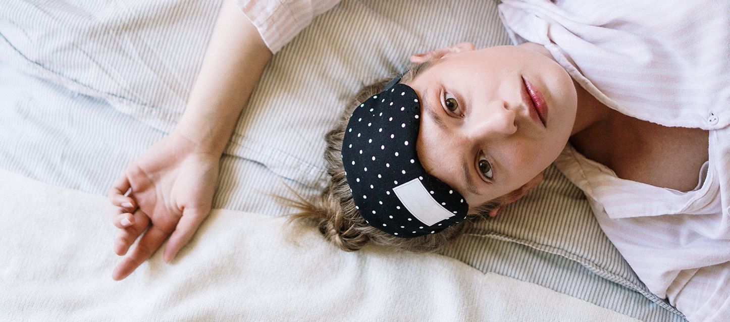 chica con pelo castaño claro, pijama a rayas blancas y rosa y antifaz en la frente, tumbada en la cama boca arriba en la cama