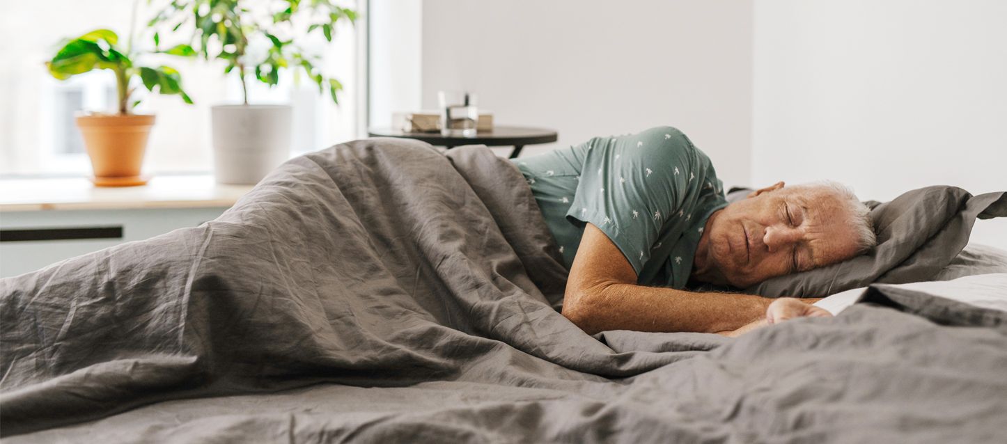 hombre mayor con pijama gris durmiendo de lado tapado con edredon gris