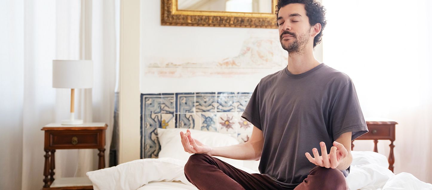 chico moreno de pelo corto con camiseta gris y pantalones granates sentado en posición de meditación en el centro de su cama
