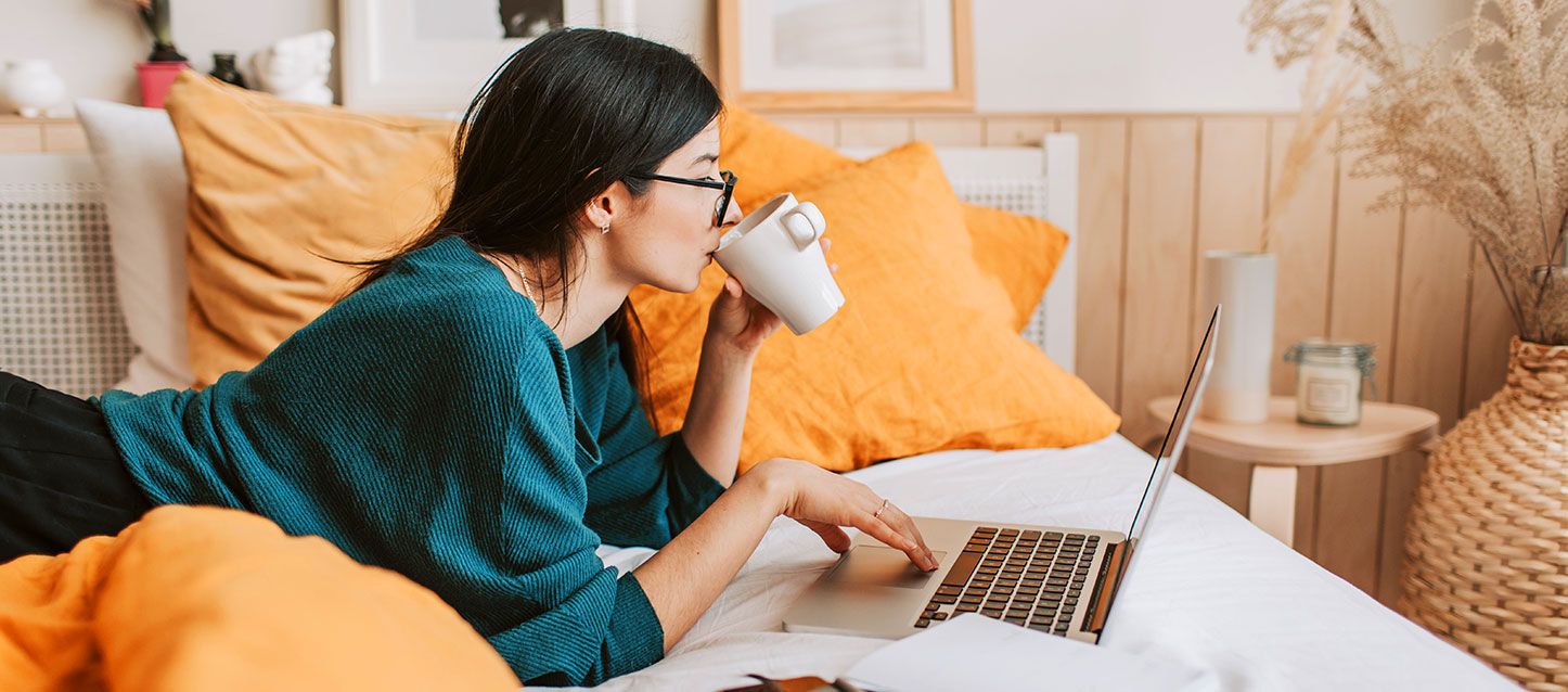 chica de pelo largo negro tumbada sobre la cama usando el ordenador portatil con una mano y con la otra tomando cafe