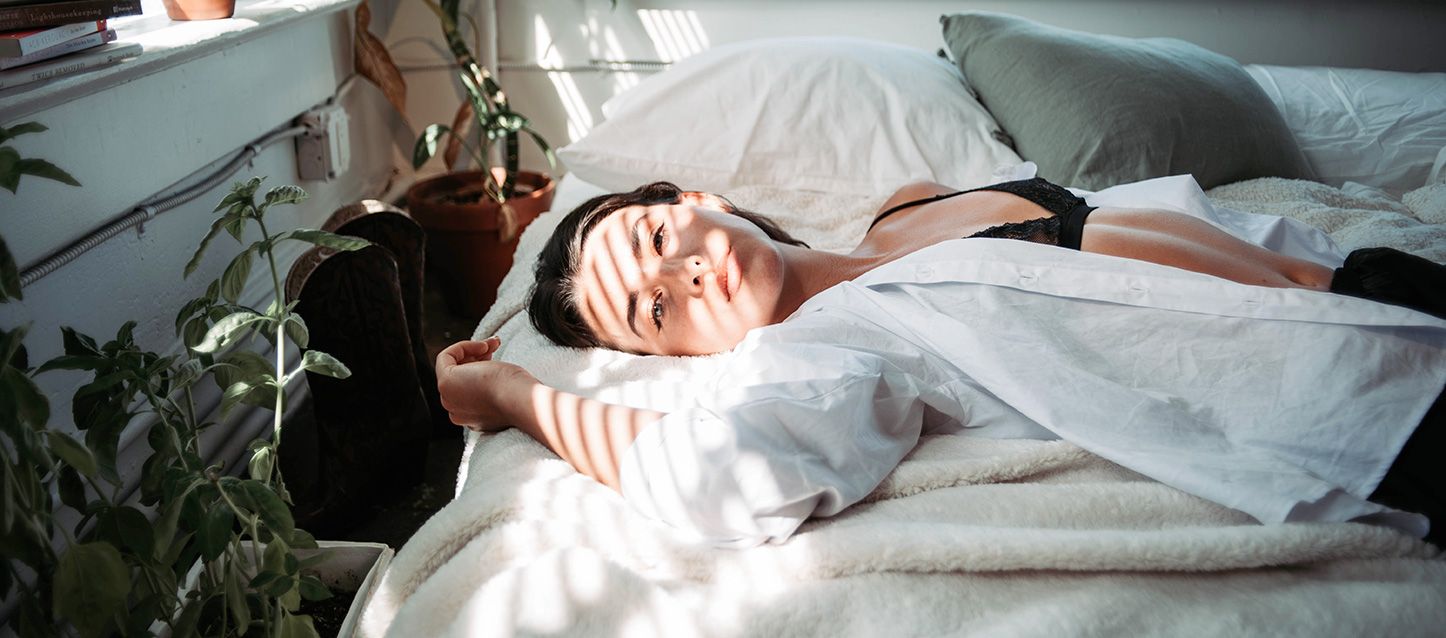 chica morena tumbada boca arriba en la cama vestida con camisa blanca
