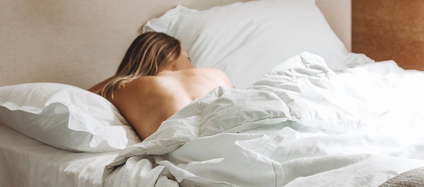 mujer rubia dormida en la cama boca abajo y tapada con el edredon hasta la espalda
