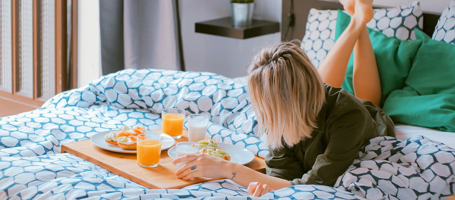 chica rubia tumbada boca abajo en la cama con las piernas flexionadas junto a una bandeja de madera con desayuno de zumo, leche y tostadas