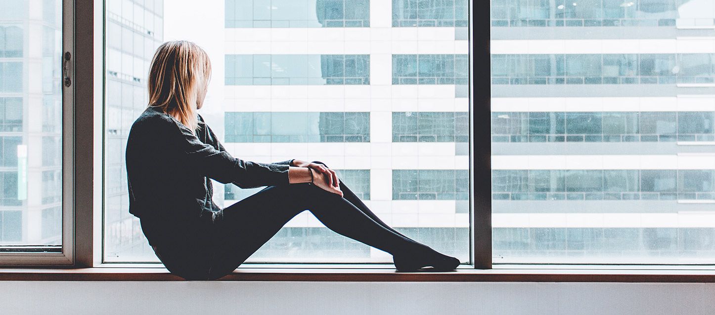 chica rubia sentada en el alfeizar de un gran ventanal con vistas al edificio de enfrente