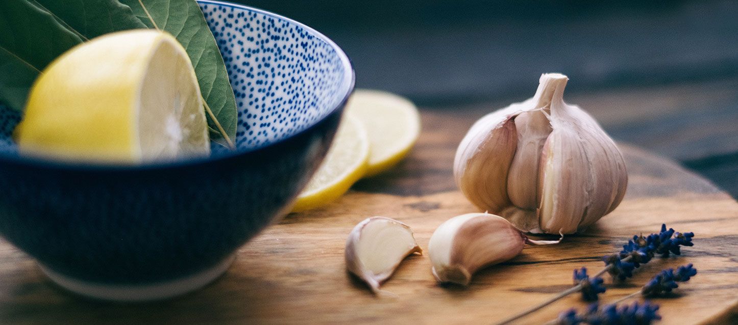 cabeza de ajo con dos dientes sobre la mesa, 2 ramas de lavanda, cuenco con limon y sus hojas y dos rodajas de limón sobre una tabla de madera