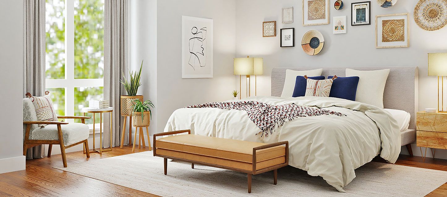 dormitorio con cama, edredon, 5 cojines, banco a los pies de la cama, cuadros decorativos en la pared, ventana con vistas a arvoles, mesita, macetas y silla