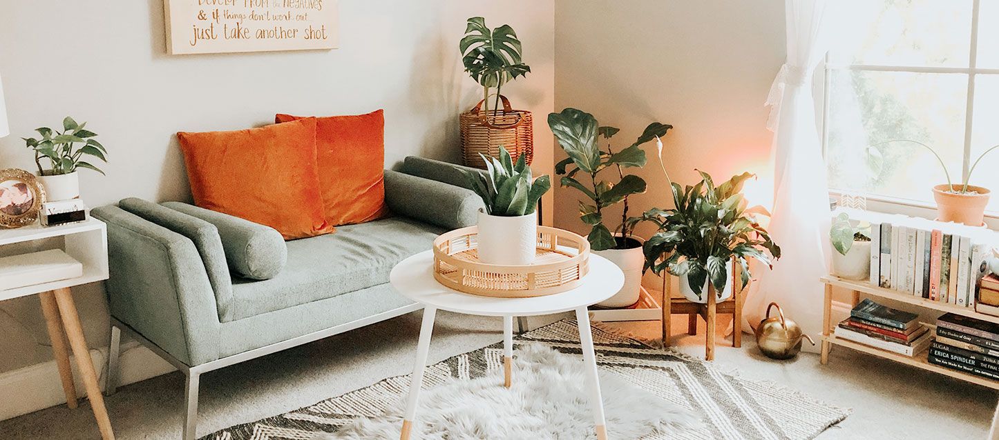 habitacion ordenada con un banco con cojines, mesita redonda blanca con una maceta, dos macetas en el suelo y estanteria con libros