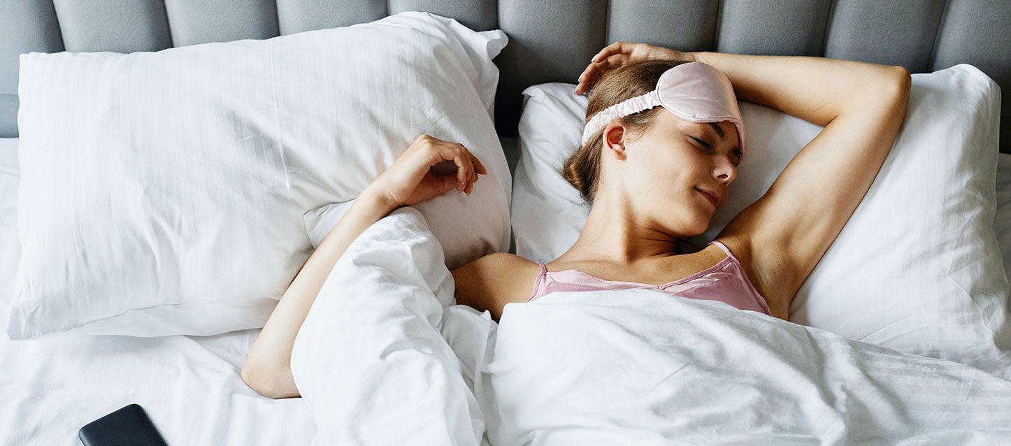 chica de pelo castaño durmiendo boca arriba metida en la cama con pijama rosa, tapada con el edredon y antifaz rosa en la frente