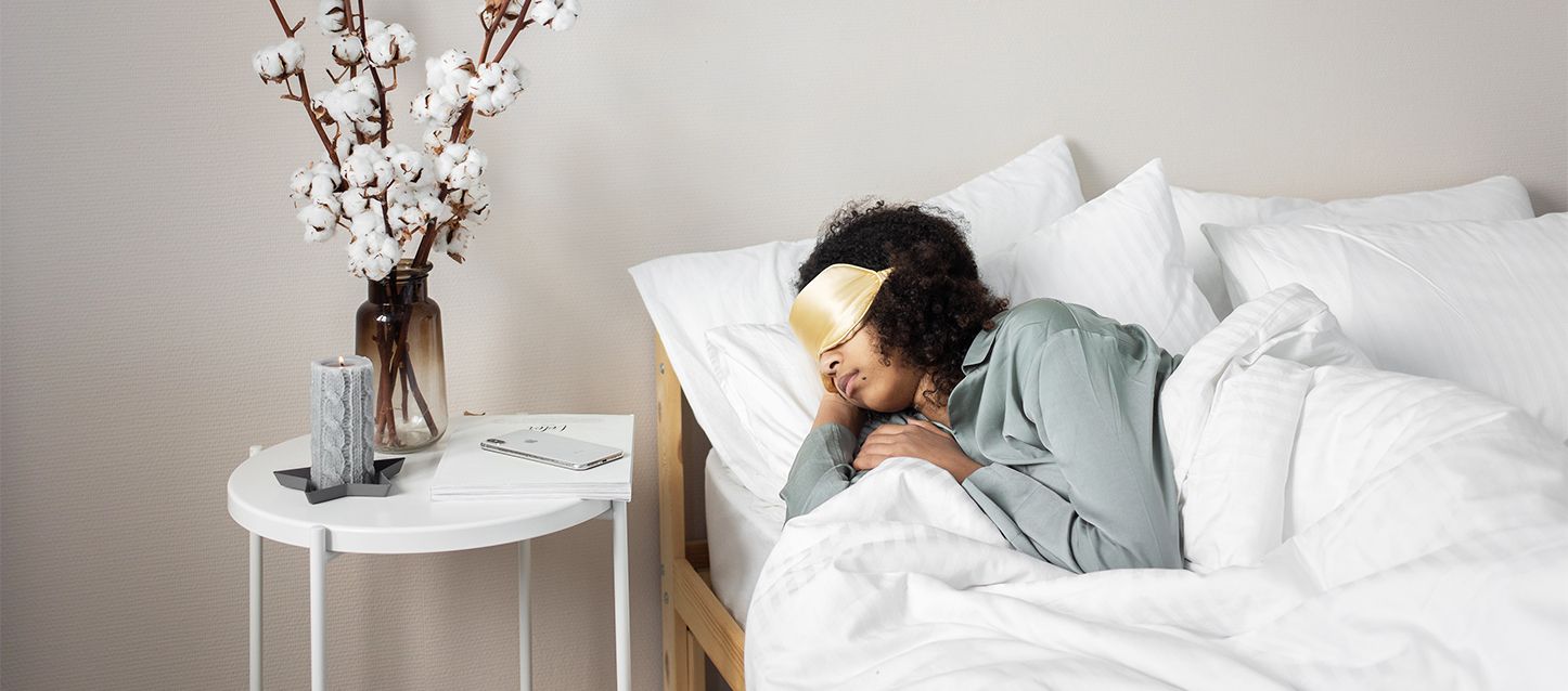 chica morena con pelo muy rizado, pijama gris y antifaz dorado durmiendo de lado junto a mesita de noche