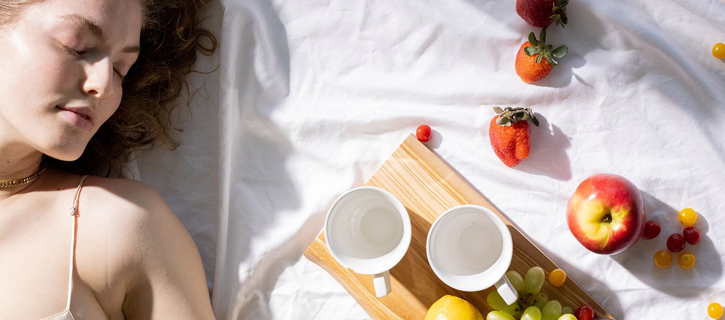 chica de pelo ondulado cataño claro, tumbada boca arriba en la cama junto a una tabla de madera con 2 tazas y varias frutas