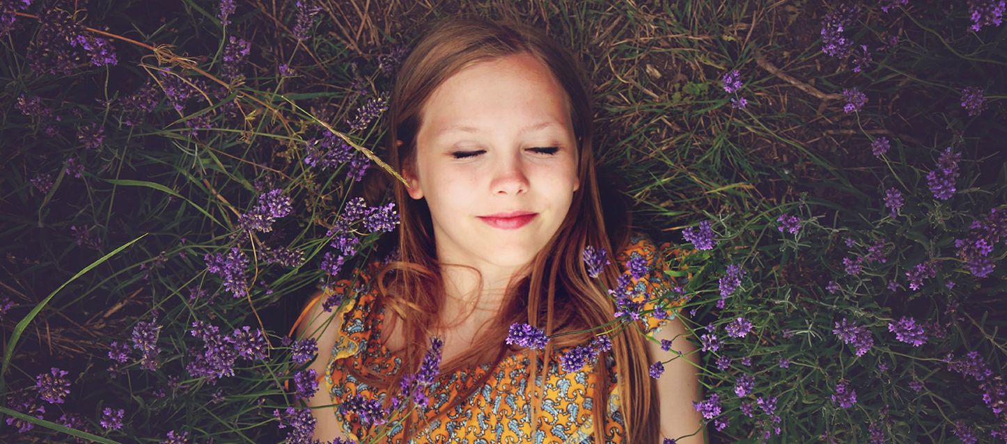 niña de pelo largo castaño con vestido naranja y dibujos blancos tumbado boca arriba en la hierba rodeada de plantas de lavanda con los ojos cerrados