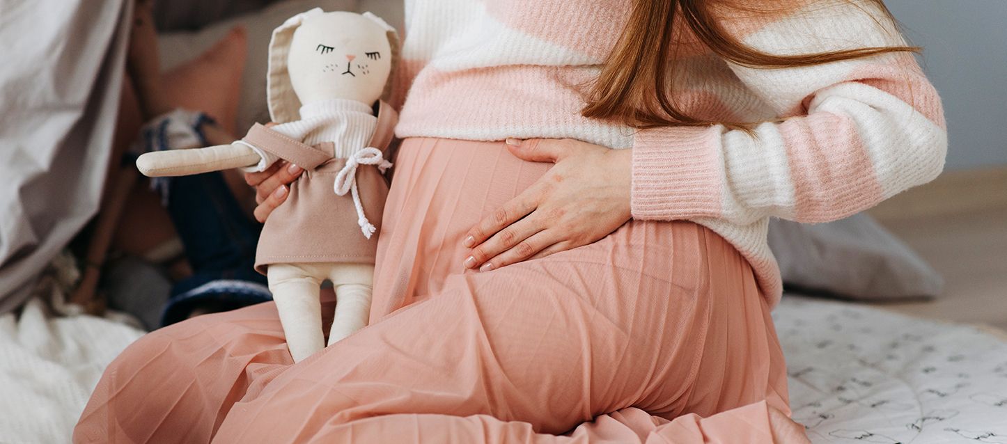 mujer embaraza sentada sobre sus piernas en la cama con falda larga rosa, jersey a rayas rosas y blancas y sujetando un conejo de peluche