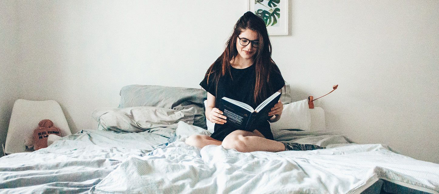 chica de pelo largo moreno sentada en la cama leyendo