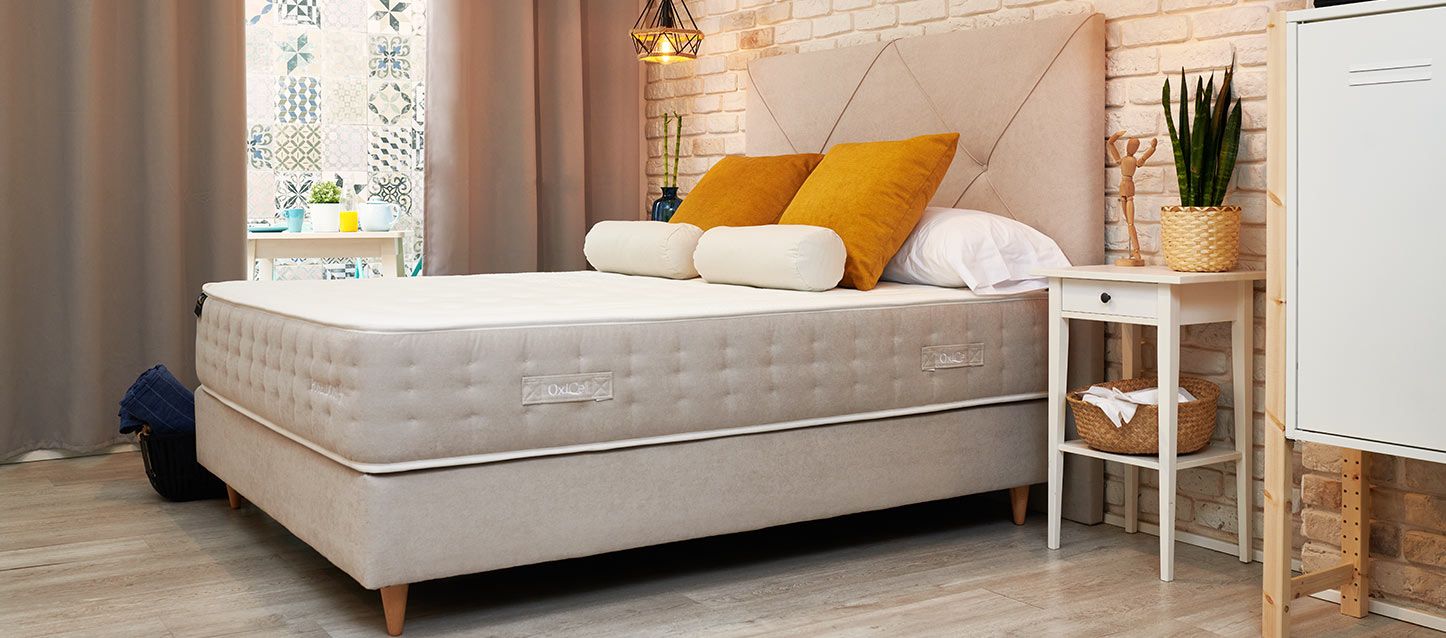 dormitorio con base tapizada gris, colchon, almohadas, 4 cojines, mesita de noche con muñeco de madera y planta