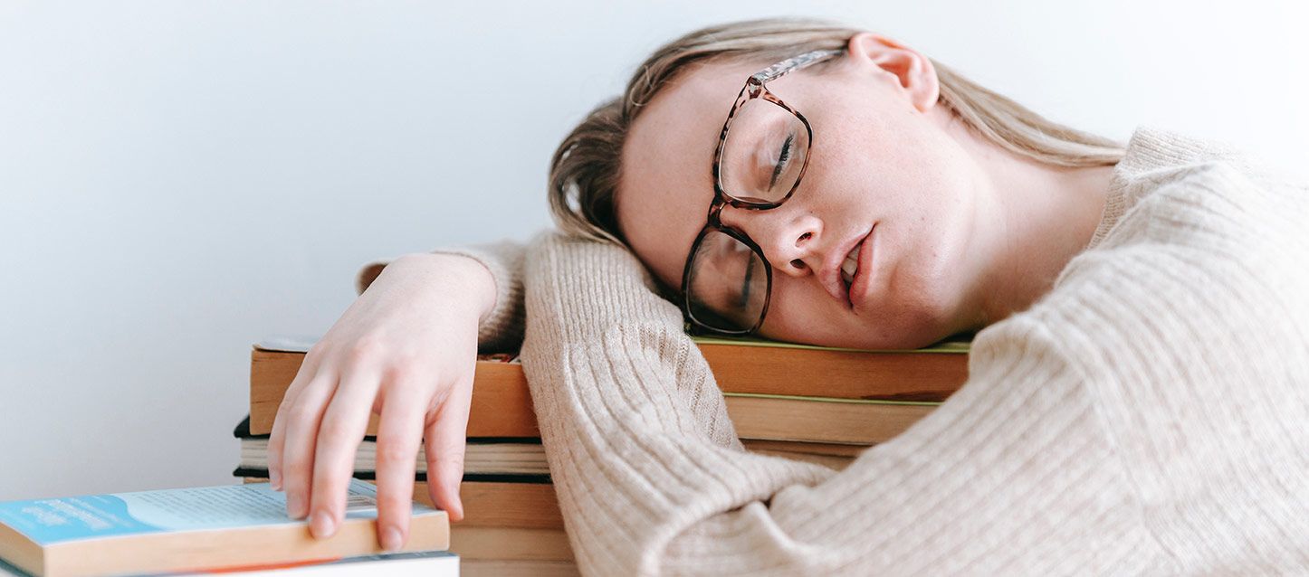 chica rubia de pelo largo con gafas durmiendo la siesta del carnero sobre unos libros apoyados en la mesa