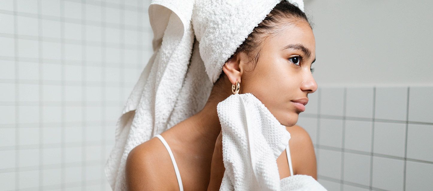 chica morena secandose al salir de la ducha con una toalla blanca enrollada en la cabeza y con otra secandose la cara