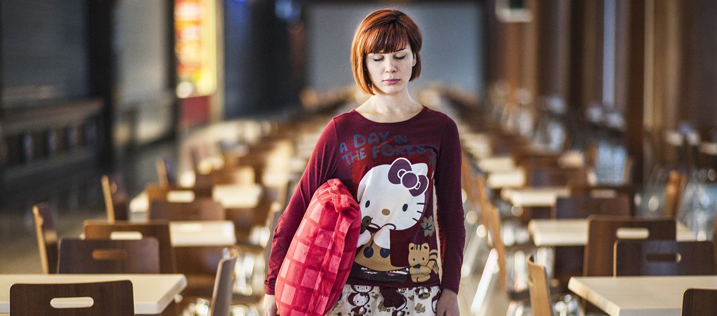 chica de media melena castaña en pijama largo con una almohada debajo de un brazo en una sala vacia