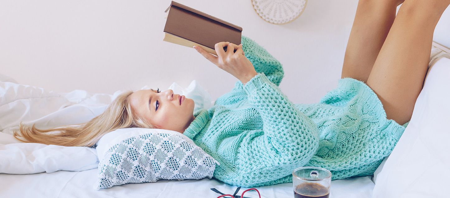 chica rubia de pelo largo tumbada boca arriba en la cama con un jersey de punto leyendo un libro, gafas y taza de cafe apoyadas sobre la cama
