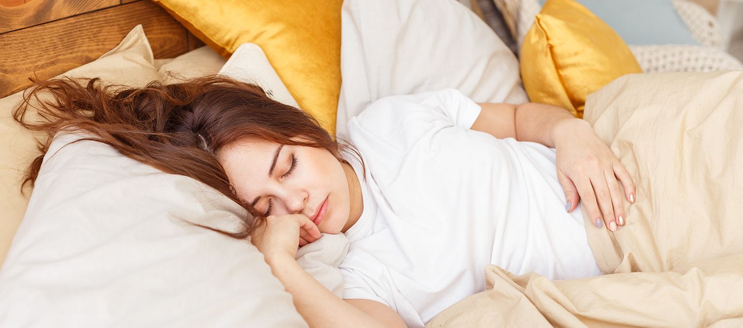 chica de pelo largo castaño, camiseta blanca durmiendo con varias almohadas y tapada con camiseta beige