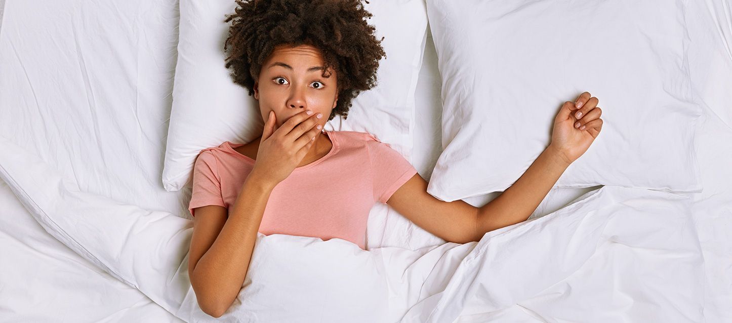chica morena de pelo rizado, pijama rosa dentro de la cama tapada con edredon blanco con la mano en la boca y gesto de sorpresa.