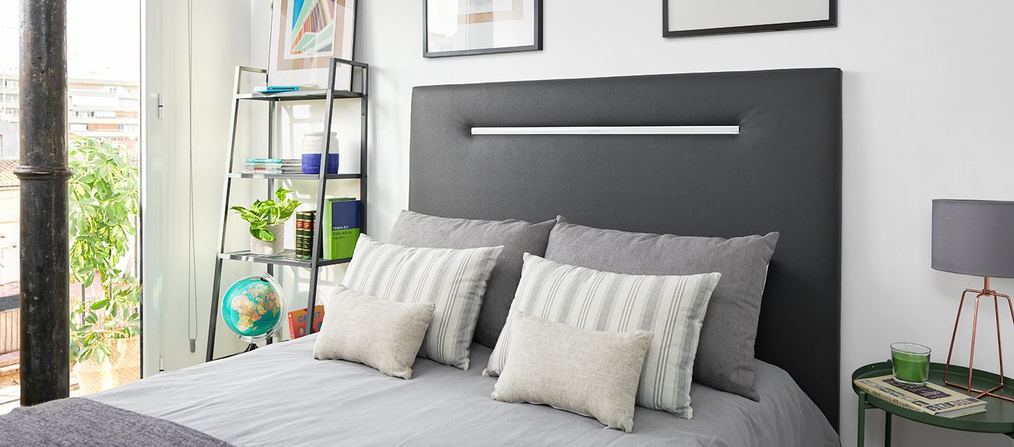 habitacion pequeña con cama, 2 almohadas y 4 cojines, estanteria con libros y mesita con una vela