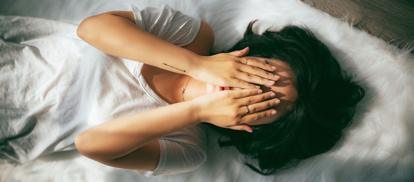 chica de pelo negro tumbada boca arriba en la cama con camiseta blanca tapandose la cara con las manos asustada por soñar que se muere