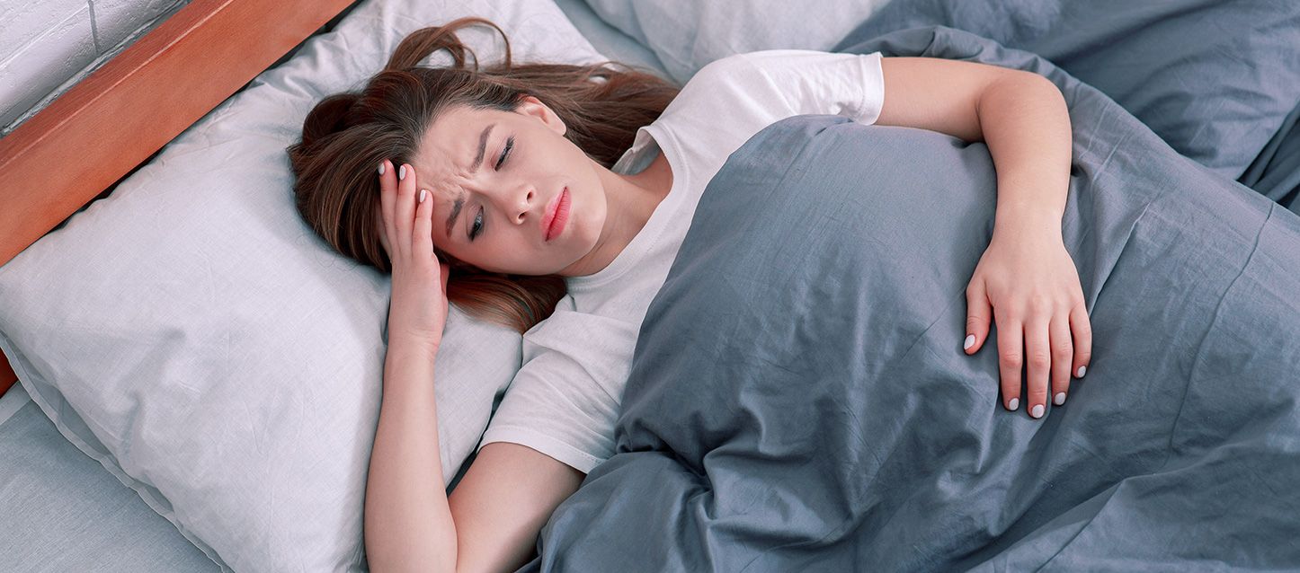 Chica metida en la cama tapada con edredon gris boca arriba despierta con la mano en la frente con gesto de malestar por soñar que se muere