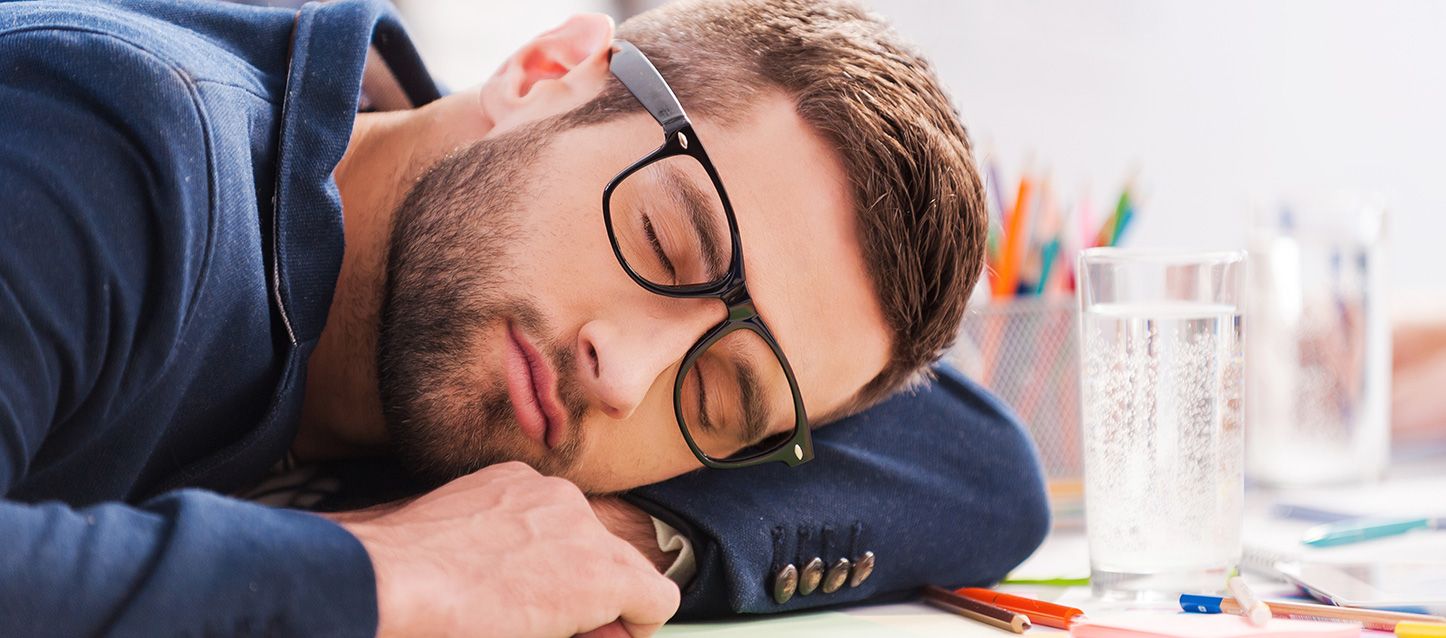 chico con barba y gafas durmiendo en una mesa junto a un vaso de agua y lapices en la mesa