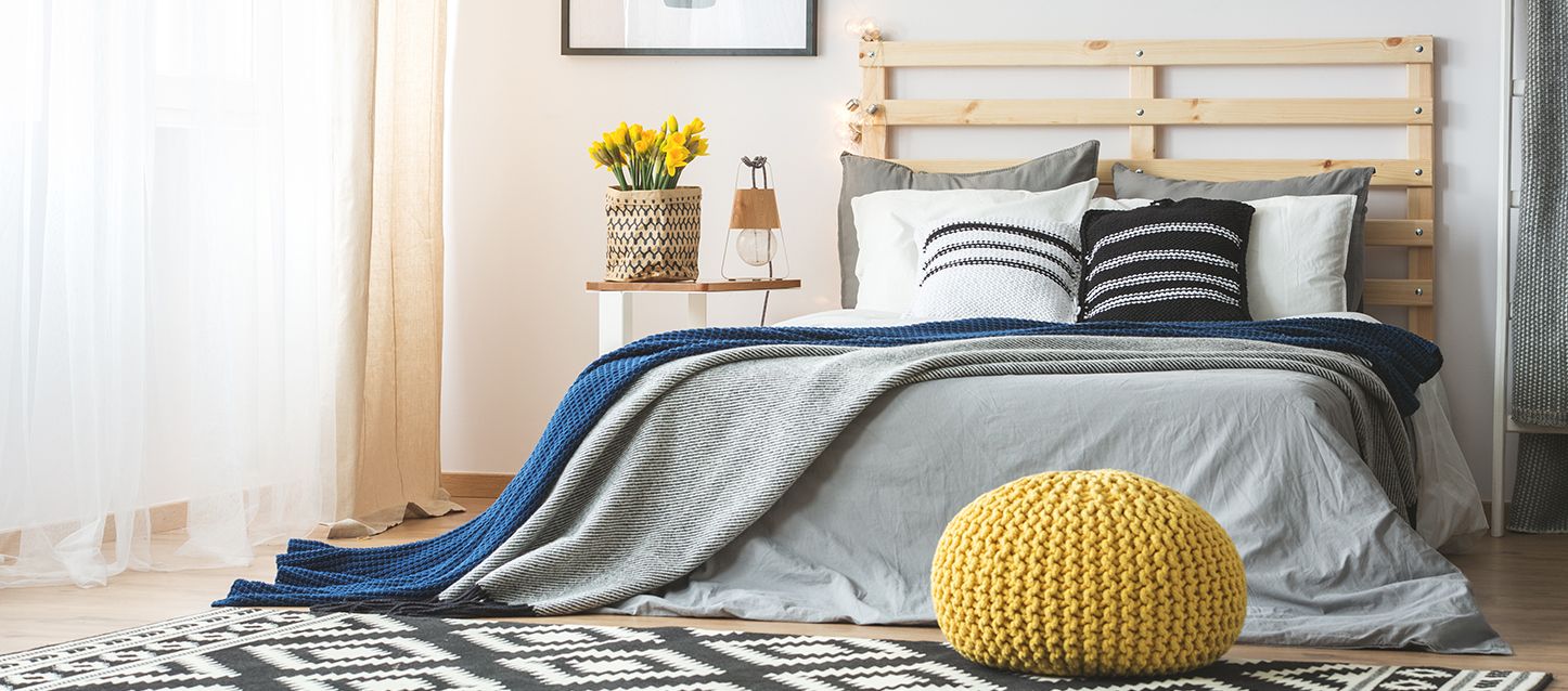 habitación con cama, mantas con lineas horizontales, cojines, cabecero de palets, mesita con lampara y maceta