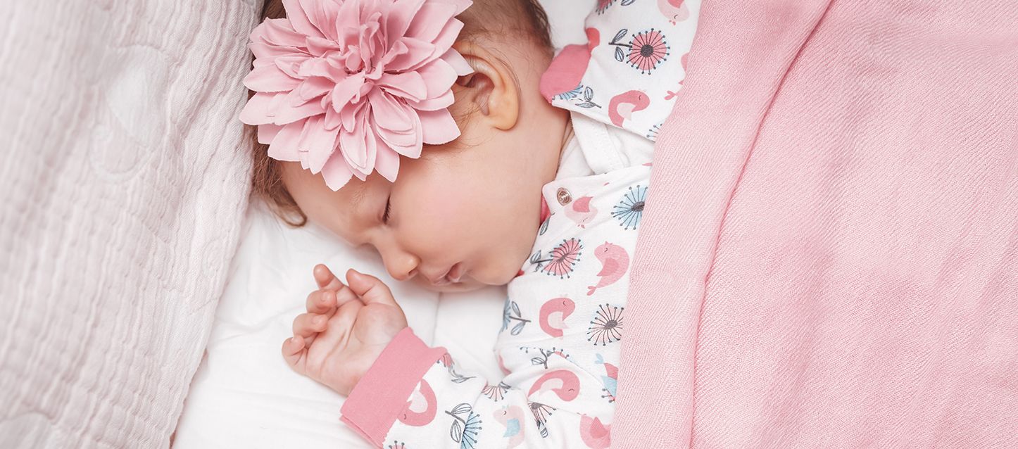bebé durmiendo boca arriba con pijama blanco y rosa, una flor decorativa en la cabeza y tapada con manta rosa