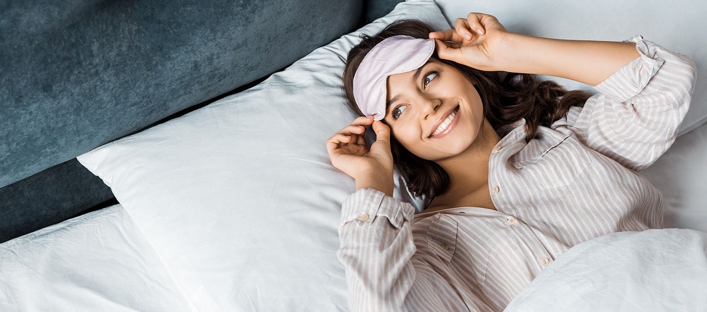 Chica de pelo moreno metida en la cama boca arriba con pijama a rayas poniéndose un antifaz
