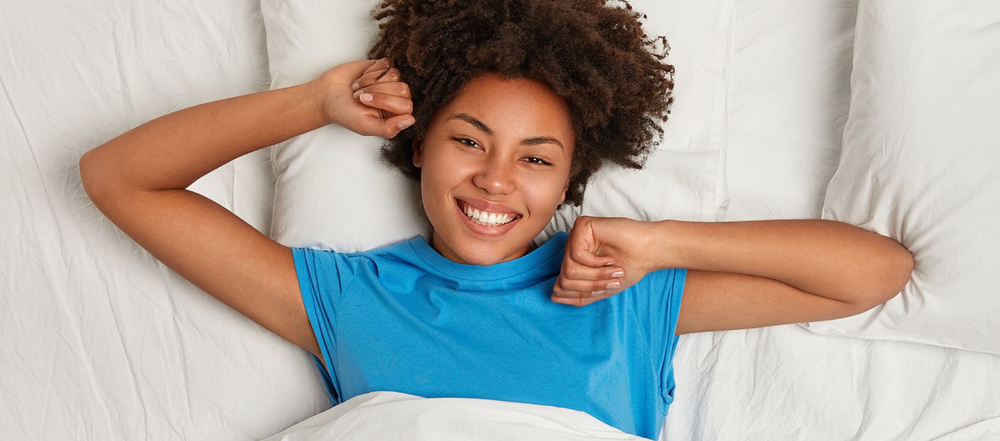 chica morena de pelo afro acostada boca arriba en la cama con camiseta azul sonriendo