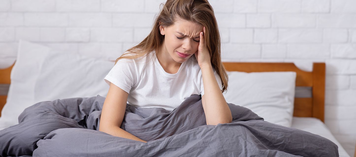 chica de pelo largo y camiseta blanca, tapada con edredon gris tocándose la frente con gesto de dolor