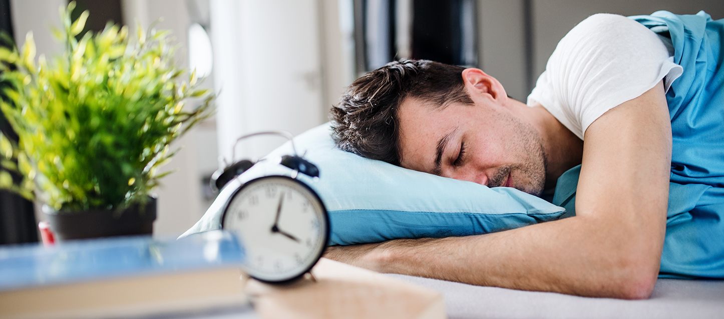 chico durmiendo boca abajo tapado con sábana azul, almohada azul claro y sobre la mesita un despertador negro, un libro y una planta
