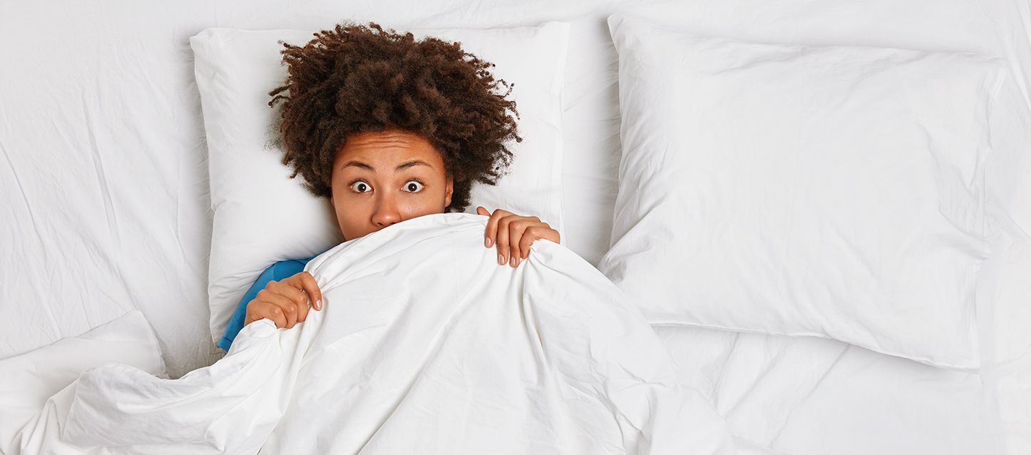 chica morena con pelo afro metida en la cama tapada con el edredon hasta la nariz con cara de asustada