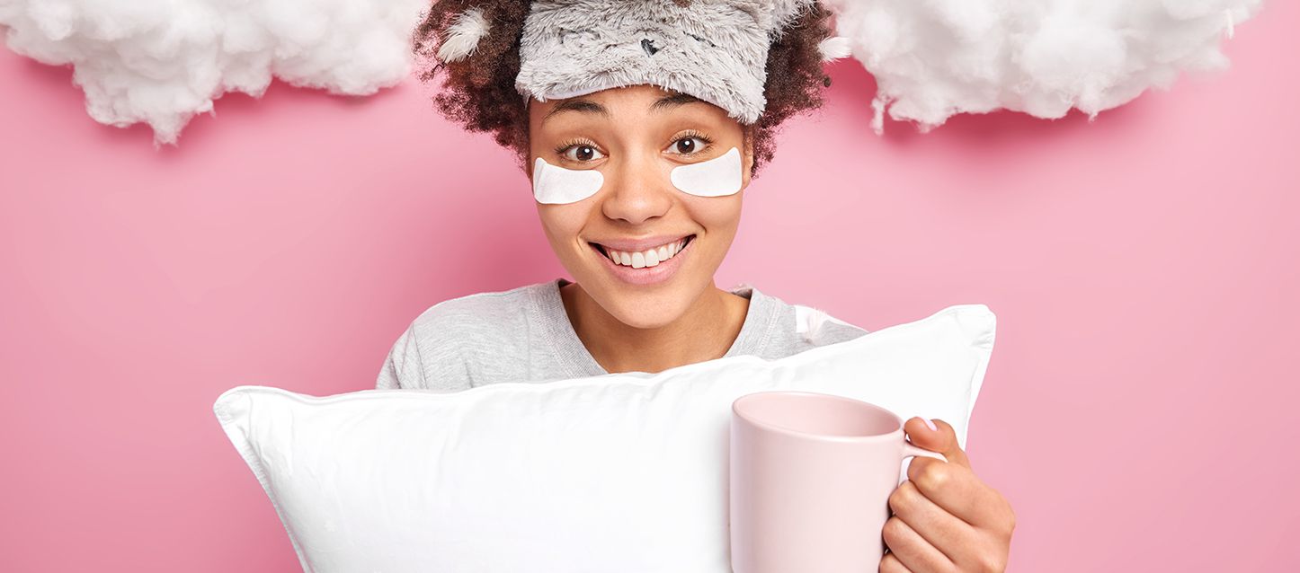 chica morena con antifaz en la cabeza sujetando una almohada y una taza rosa