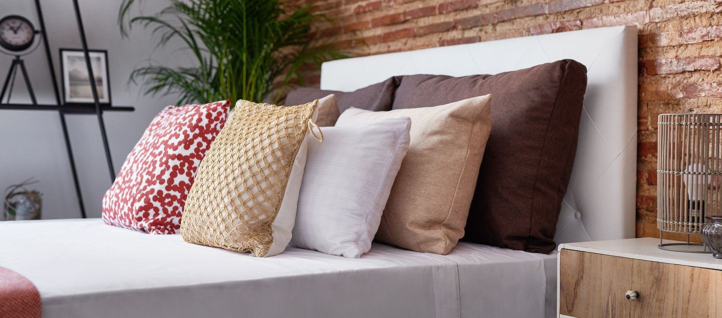 Cabecero de polipiel blanco, 8 cojines de diferentes colores colocadas dos a dos sobre la cama con sábana gris