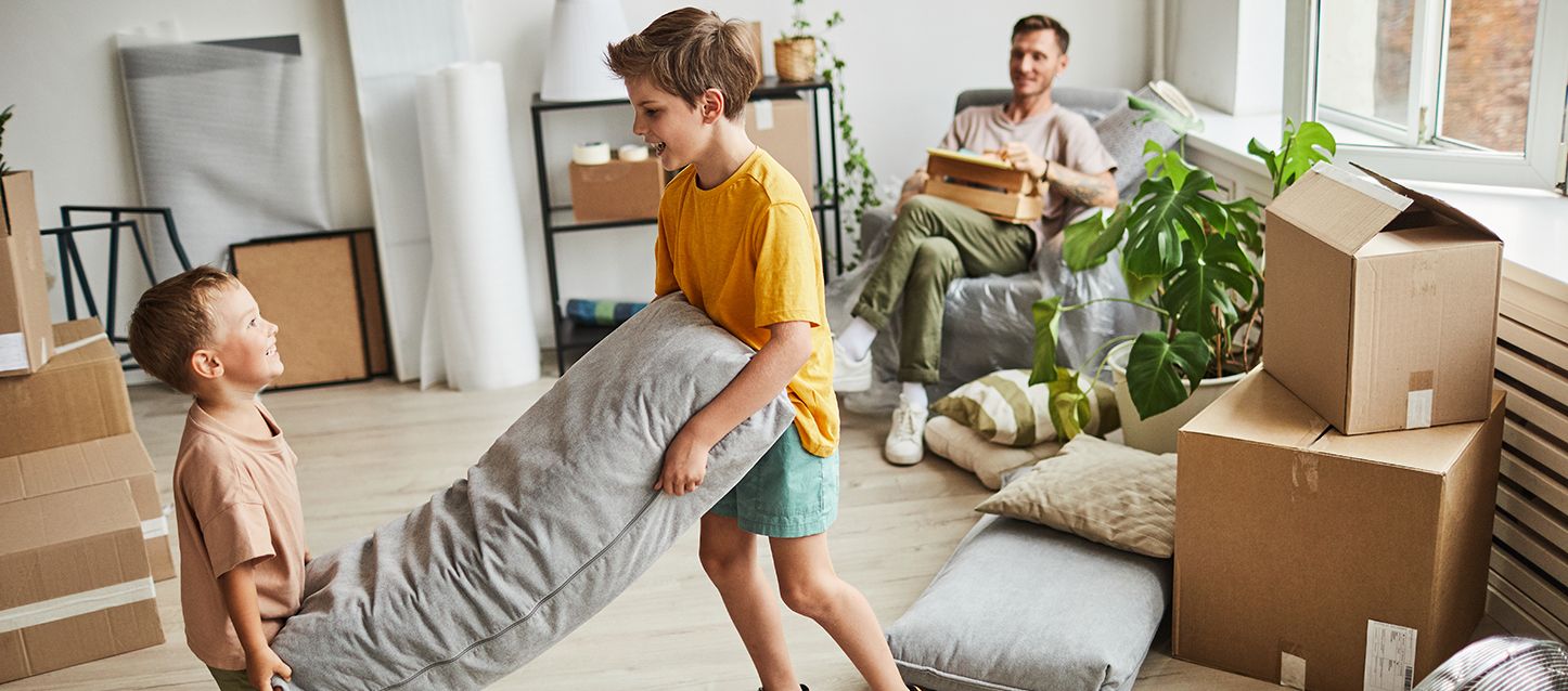 dos hermanos jugando con una almohada en una casa de mudanza con el padre mirando