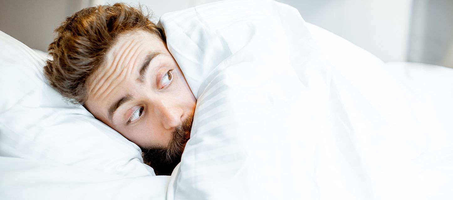 hombre con barba metido en la cama tapado con el edredón blanco hasta la cara con cara de asustado