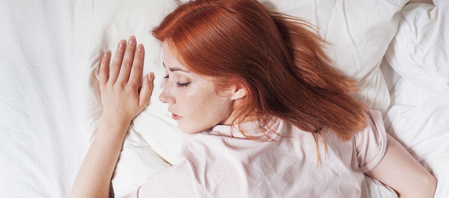 Chica pelirroja dormida boca abajo con camiseta de pijama rosa palo con la cabeza ladeada a la izquierda
