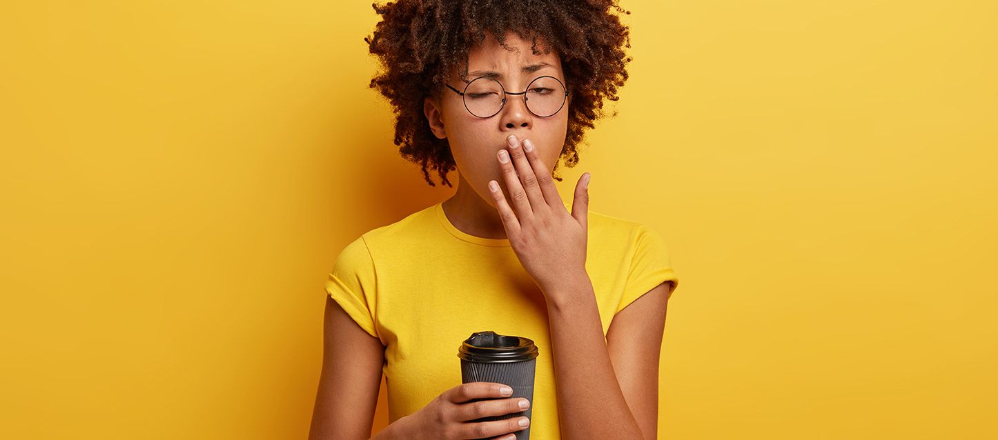 Chica morea con camiseta amarilla con una café en la mano y bostezando