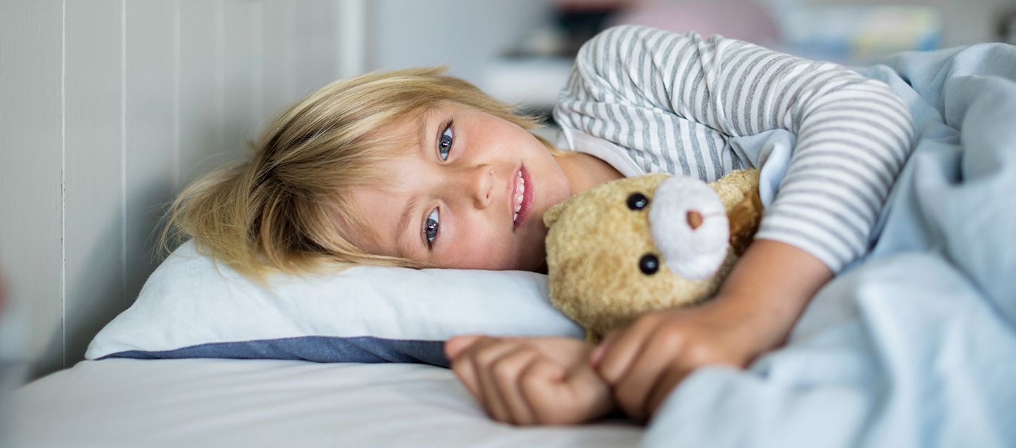 niño pequeño rubia de ojos verdes acostado de lado en la cama abrazando a un osito de peluche mirando hacia la camara