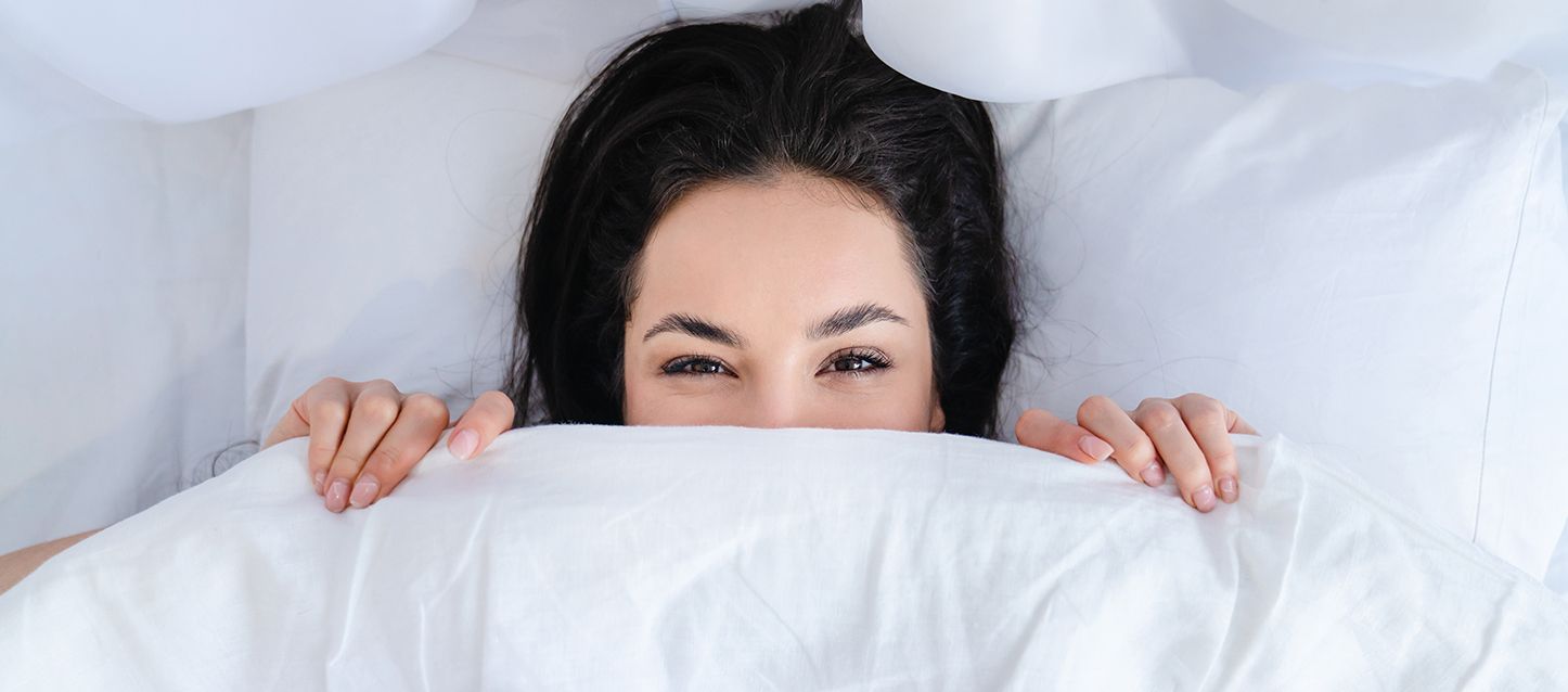 Chica morena de pelo largo metida en la cama tapada con el edredon hasta la nariz