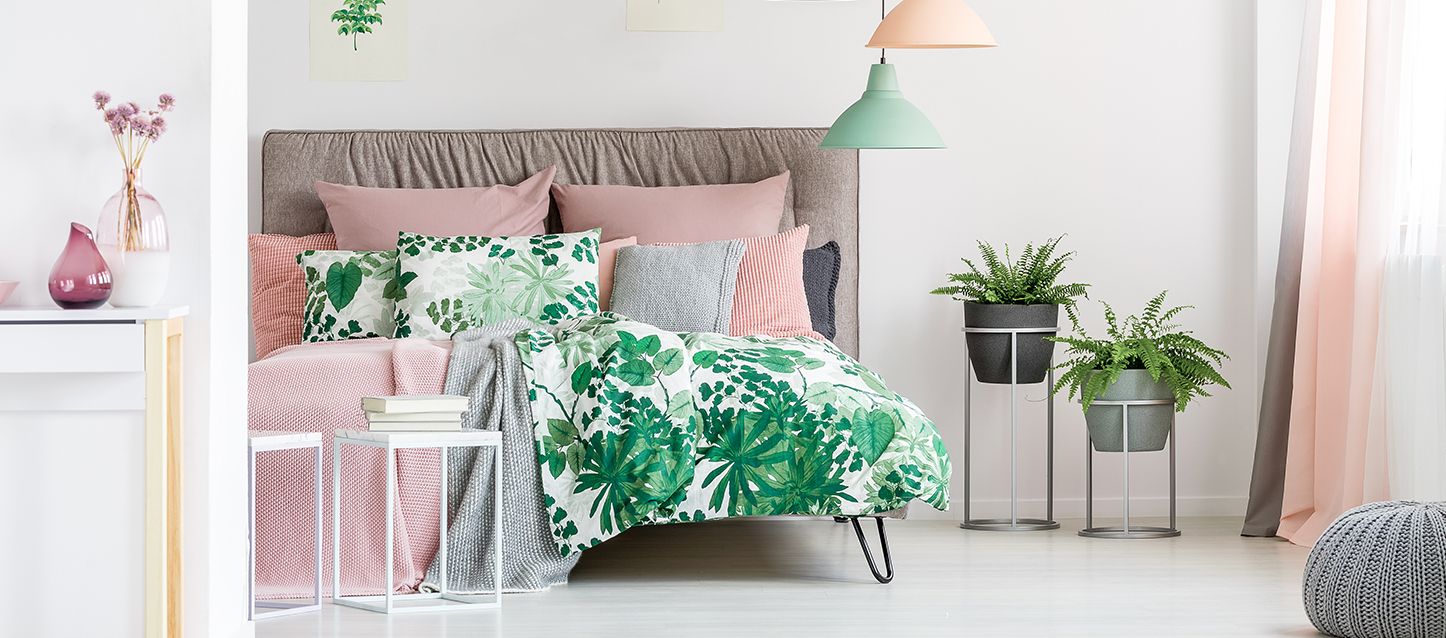 Dormitorio con cama, edredón, cojines, 2 macetas y dos mesitas, una con libros