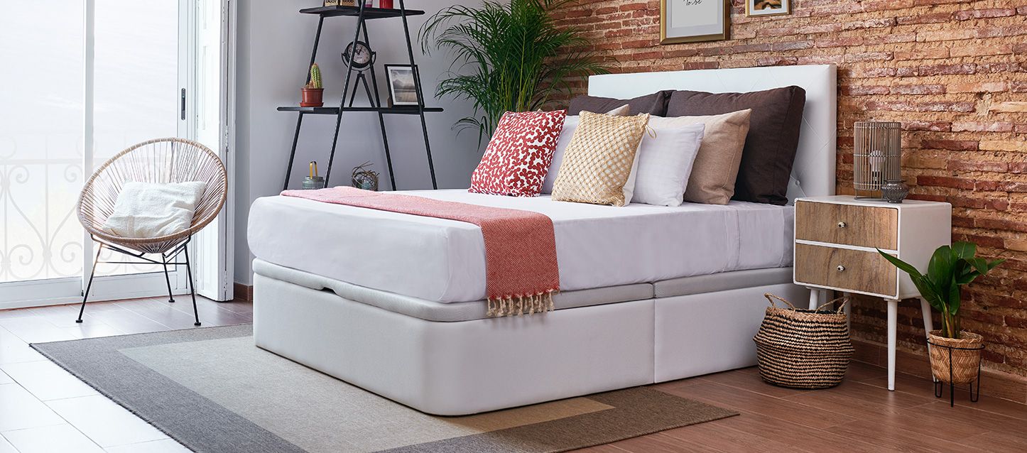 Dormitorio con canape, colchon, cojines, almohadas, mesita de noche con lámpara, silla y estanteria con objetos decorativos