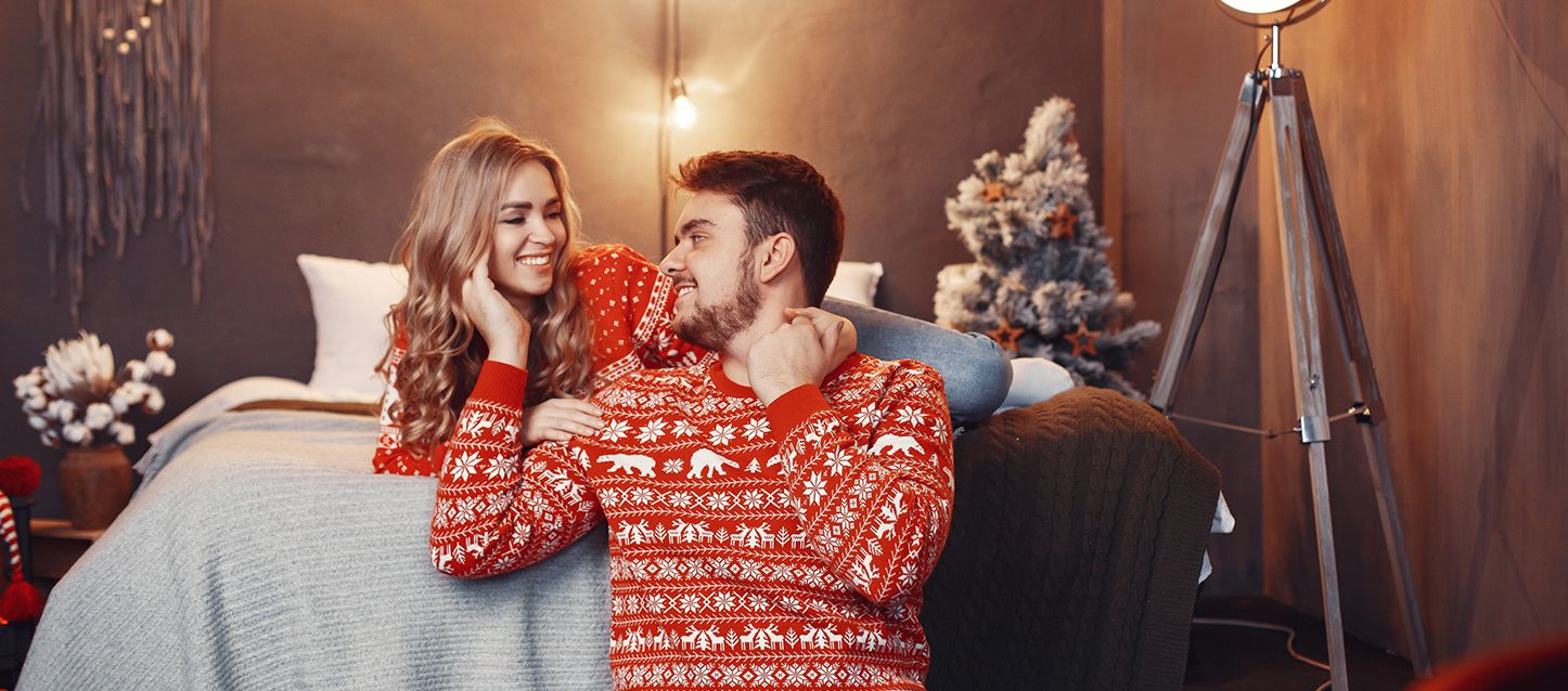 Pareja heterosexual con jerseys navideños apoyados en una cama mirándose felices