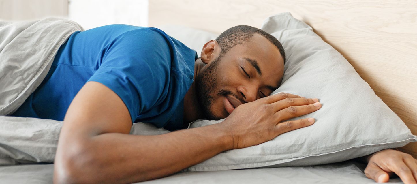 Hombre tumbado boca abajo durmiendo plácidamente con una mano sobre la almohada gris y la otra por debajo