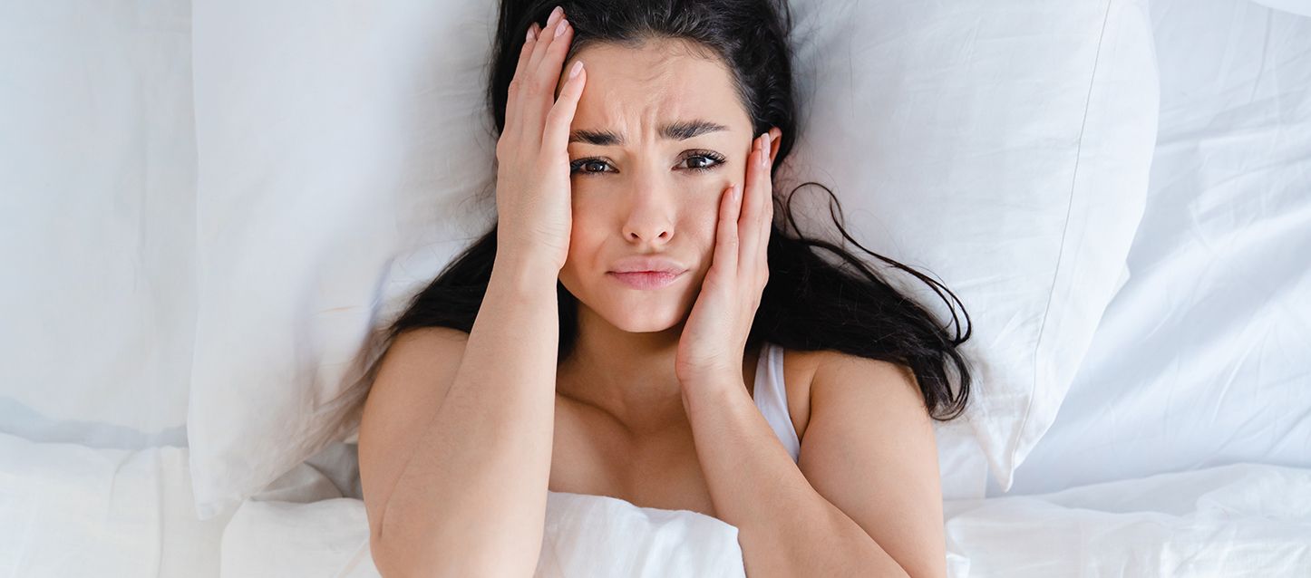 Chica de pelo largo negro metida en la cama cogiendo su cara con las dos manos con cara de asustada