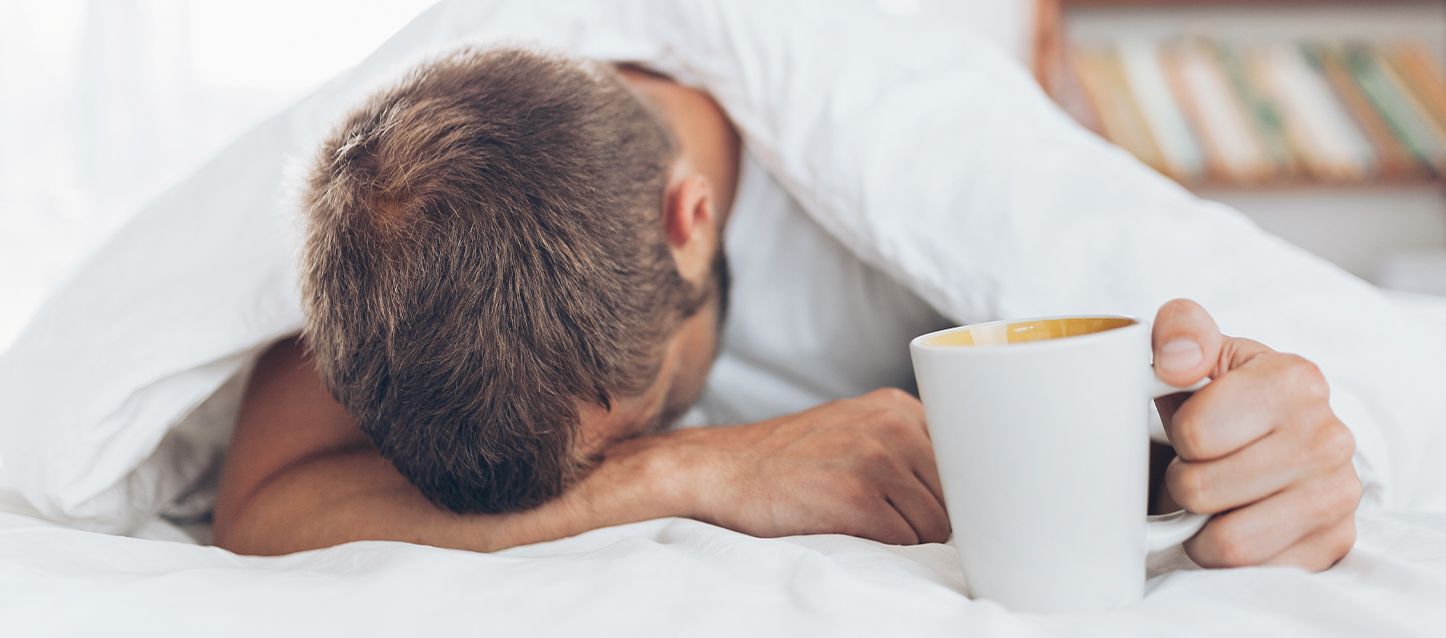 Hombre en la cama boca abajo cansado y con malestar sujetando una taza blanca en la mano